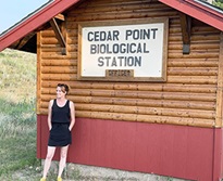 A photo of Sarah Kaizar infront of a small cabin with a sign that says 'Cedar Point Biological Station.'